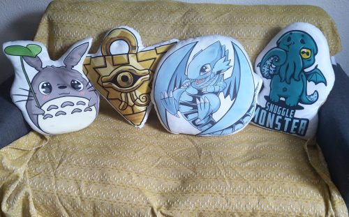 A photo of a couch with four shaped pillows: Totoro, Millenium Puzzle, Blue Eyes White Dragon, and a pillow showing a chibi-style Cthulhu rubbing its eyes called "Snuggle Monster"